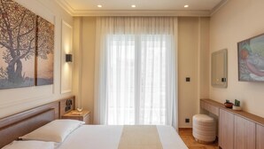 A hotel room with a large bed, a bedside table with a lamp, a small round ottoman, a wooden dresser, and a large window with sheer curtains. - Mixalakopoulou Luxury Apartment (Athens)