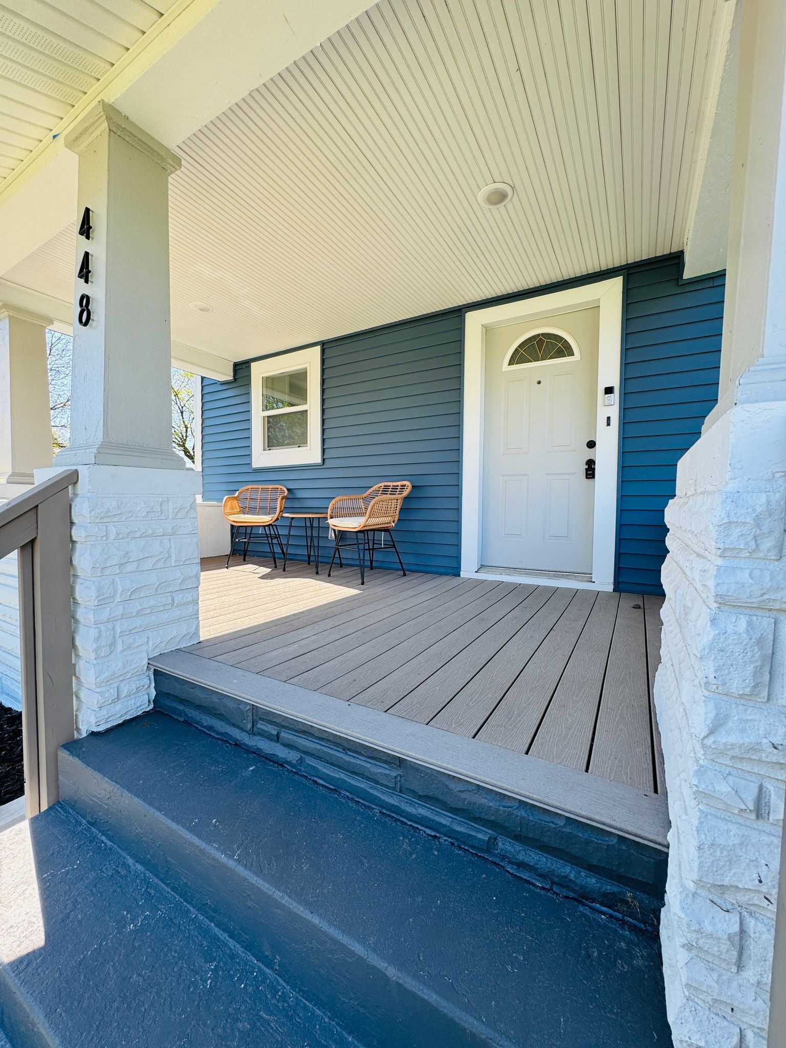 A covered porch with a wooden deck, two wicker chairs, a white door, and a mailbox.