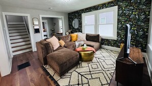 Un salón con un sofá seccional marrón, una mesa de centro y un mueble para televisor. Se ve una escalera que lleva a otra habitación, y una ventana con persianas.
