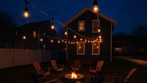 Un patio trasero con una fogata rodeada de sillas Adirondack, luces colgantes y una casa al fondo.