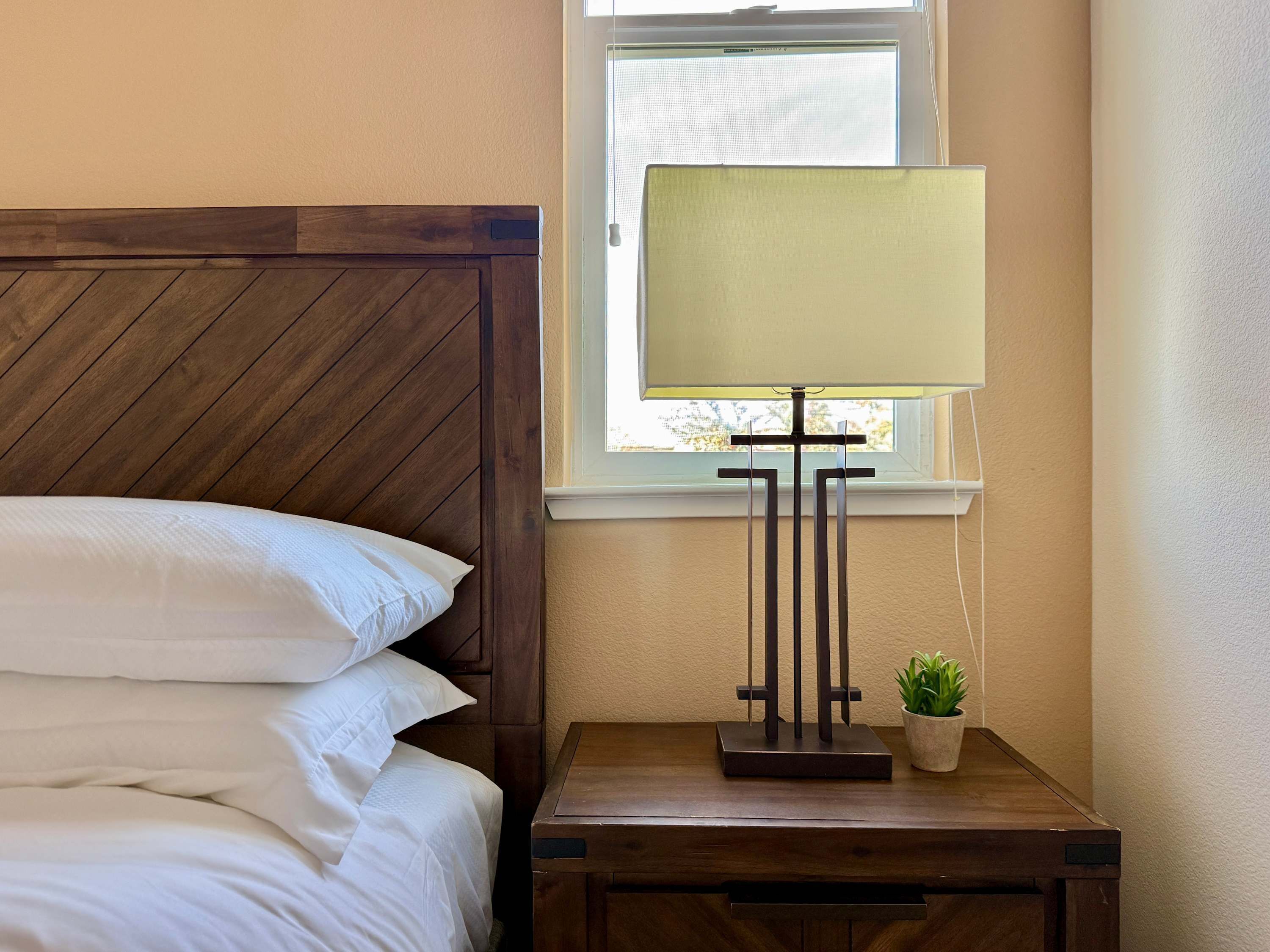 A neatly made bed with white linens, a wooden headboard, a bedside table with a lamp, and a small potted plant.