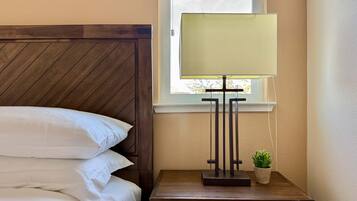 A neatly made bed with white linens, a wooden headboard, a bedside table with a lamp, and a small potted plant.