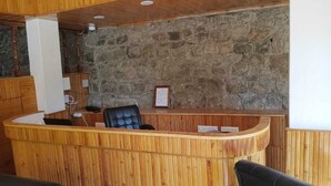 A reception area with a wooden reception desk, a black leather chair, and a stone wall behind. - Tirthan Villa Resort (Banjar)