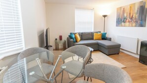 A modern living room with a grey sectional sofa, glass dining table, and chairs, a floor lamp, and a cityscape painting on the wall. - Bedford Meadows (Bedford)