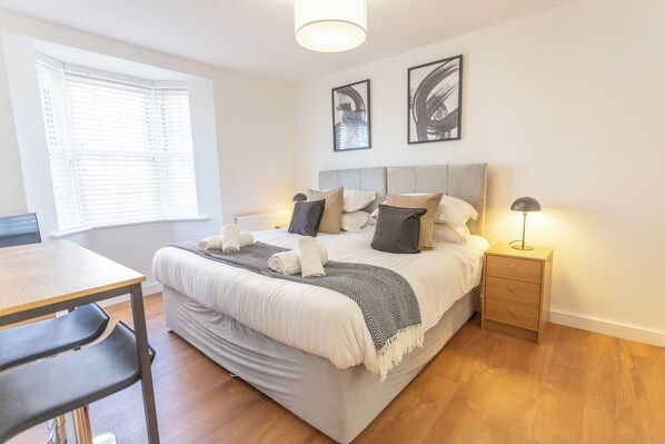 A neatly arranged bedroom with a bed, bedside table, lamp, and a window with blinds.