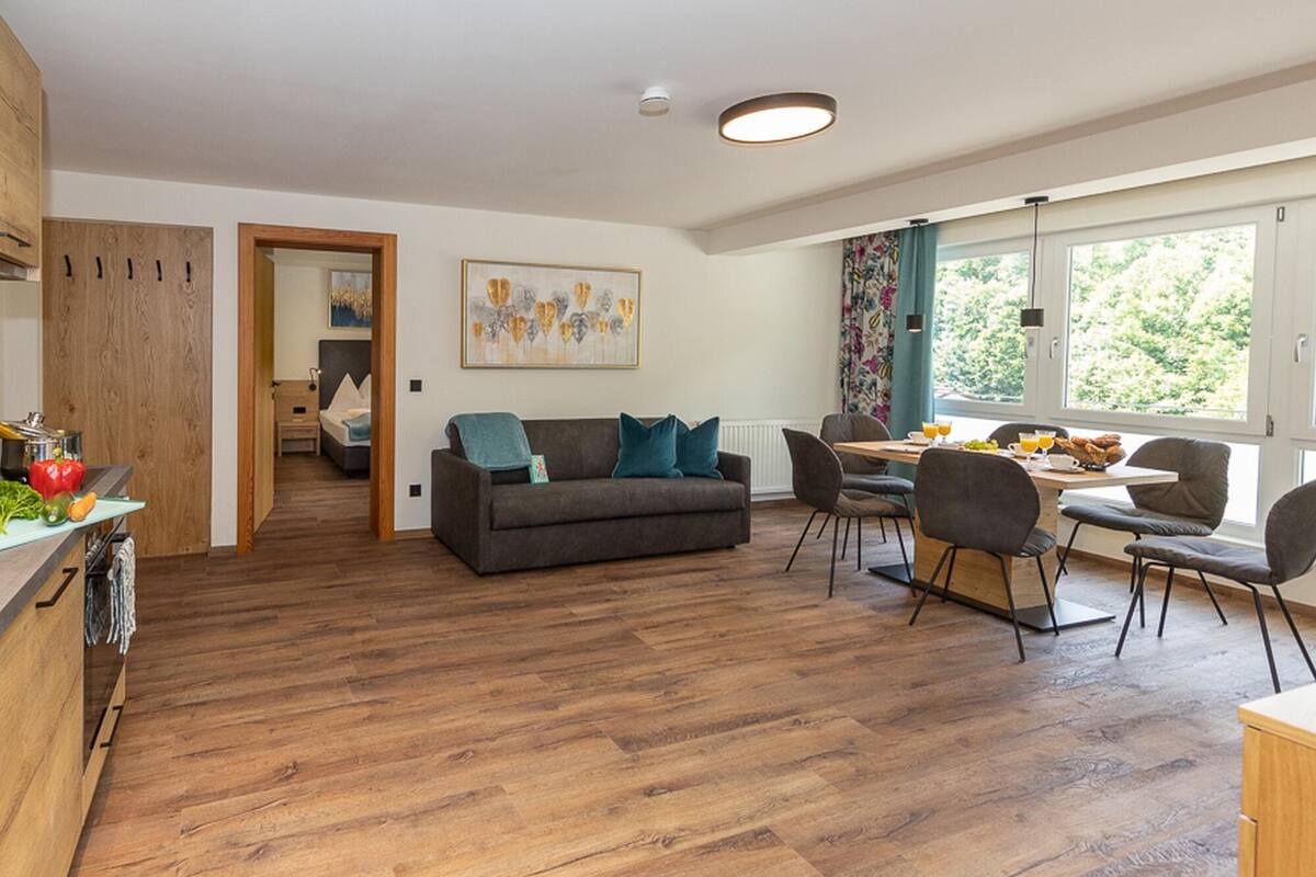 Eine moderne Küche mit Holzschränken, ein Essbereich mit Tisch und Stühlen sowie ein Wohnzimmer mit Sofa und einem Gemälde.