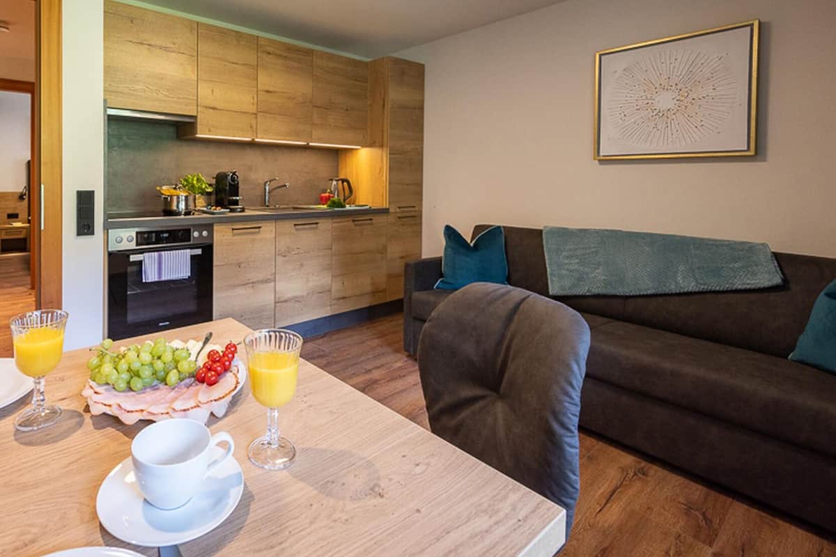 Eine moderne Küche mit Holzausstattung, einem Esstisch mit Tellern, Gläsern und einer Obstplatte. Im Wohnbereich steht ein braunes Sofa mit blauen Kissen.