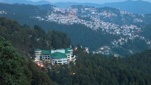 A hotel with multiple green-roofed buildings nestled in a forested valley, surrounded by a densely populated town. - Hotel CK International By Pax Hotels (Shimla)