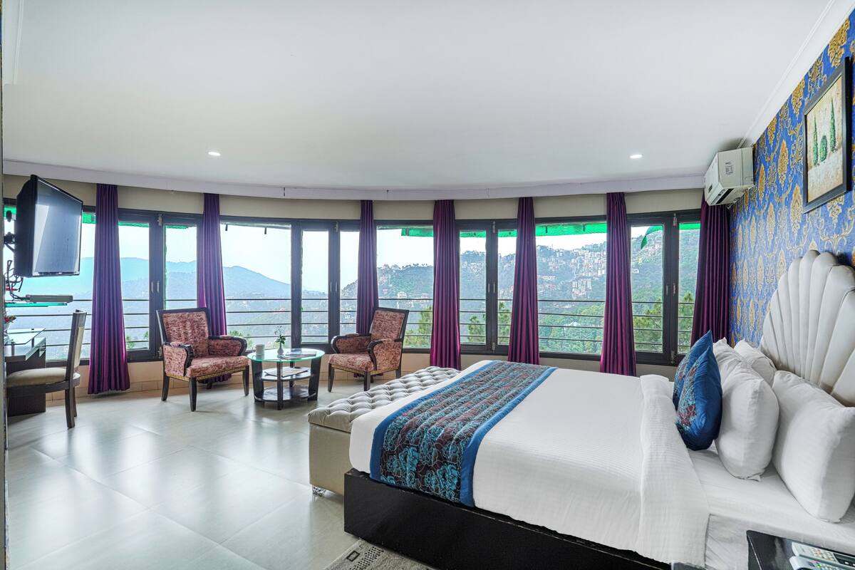 Habitación de hotel con una cama grande, dos sillones, una mesita y un televisor. La habitación tiene ventanas amplias con cortinas moradas y vistas a un paisaje montañoso.