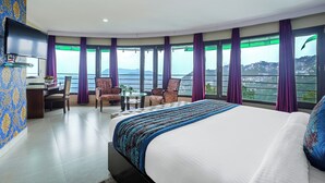 A hotel room with a large bed, two armchairs, a desk, and a TV. The room has large windows with purple curtains and a view of the city. - Hotel CK International By Pax Hotels (Shimla)