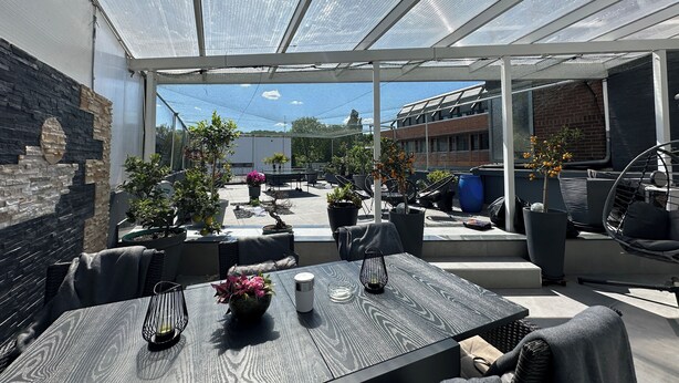 Un patio coperto con mobili da esterno, piante in vaso e vista sugli edifici circostanti.