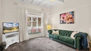 Un salon avec une télévision, un canapé vert, une fenêtre avec des rideaux, un tableau et une lampe.