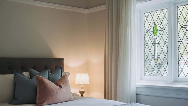 Une chambre à coucher avec un lit, des oreillers, une table de chevet, une lampe et une fenêtre ornée d’un motif géométrique.