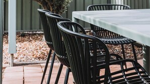 Un patio couvert avec une table et des chaises, un grand buisson et, au fond, une clôture en métal.