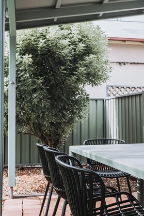 A covered patio area with a table and chairs, a large bush, and a metal fence in the background. - Boleyn | Timeless Charm (Orange)