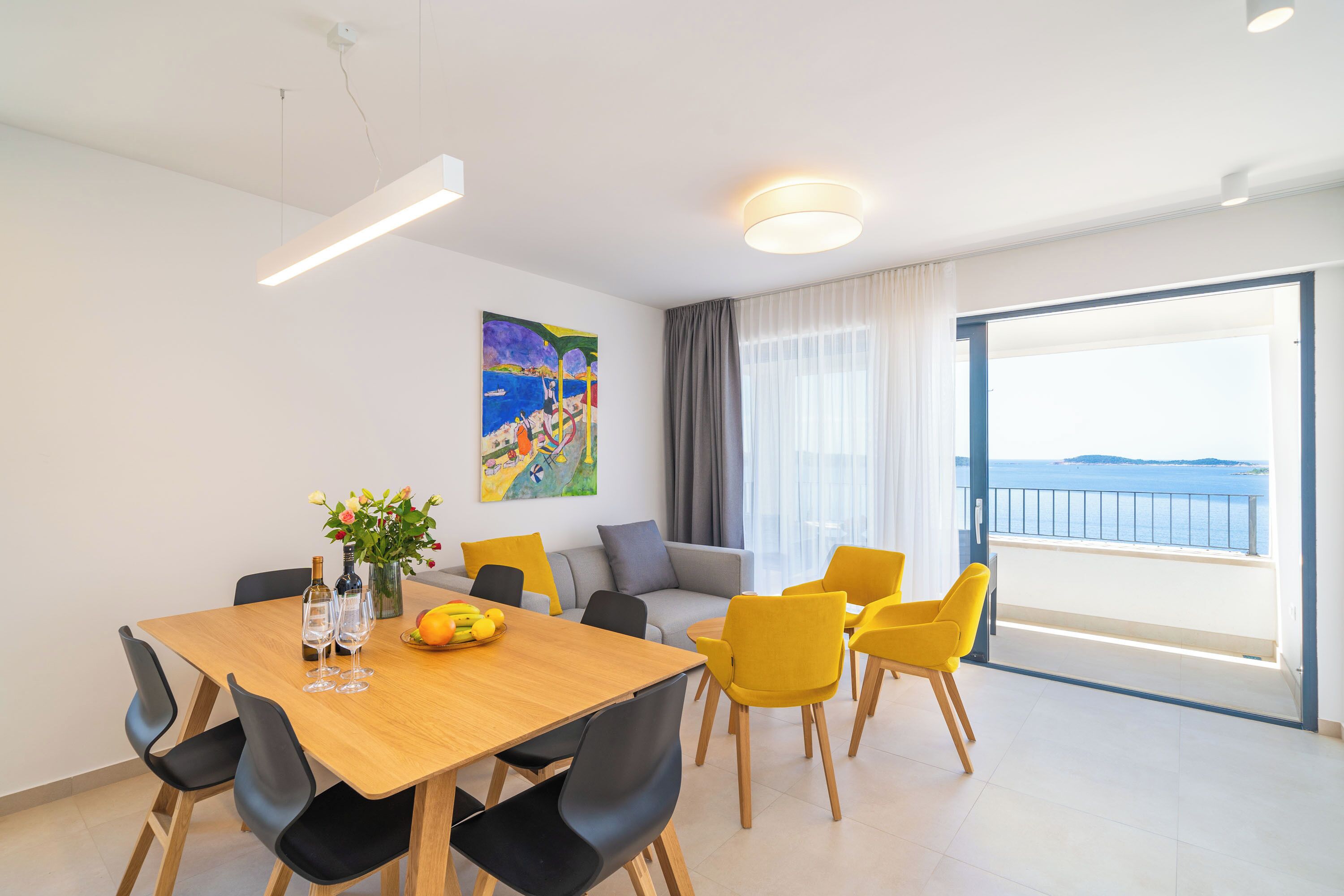 A modern living room with a dining area, featuring a large window with a view of the sea, a painting on the wall, and a sofa with yellow chairs.