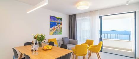 A modern living room with a dining area, featuring a large window with a view of the sea, a painting on the wall, and a sofa with yellow chairs.