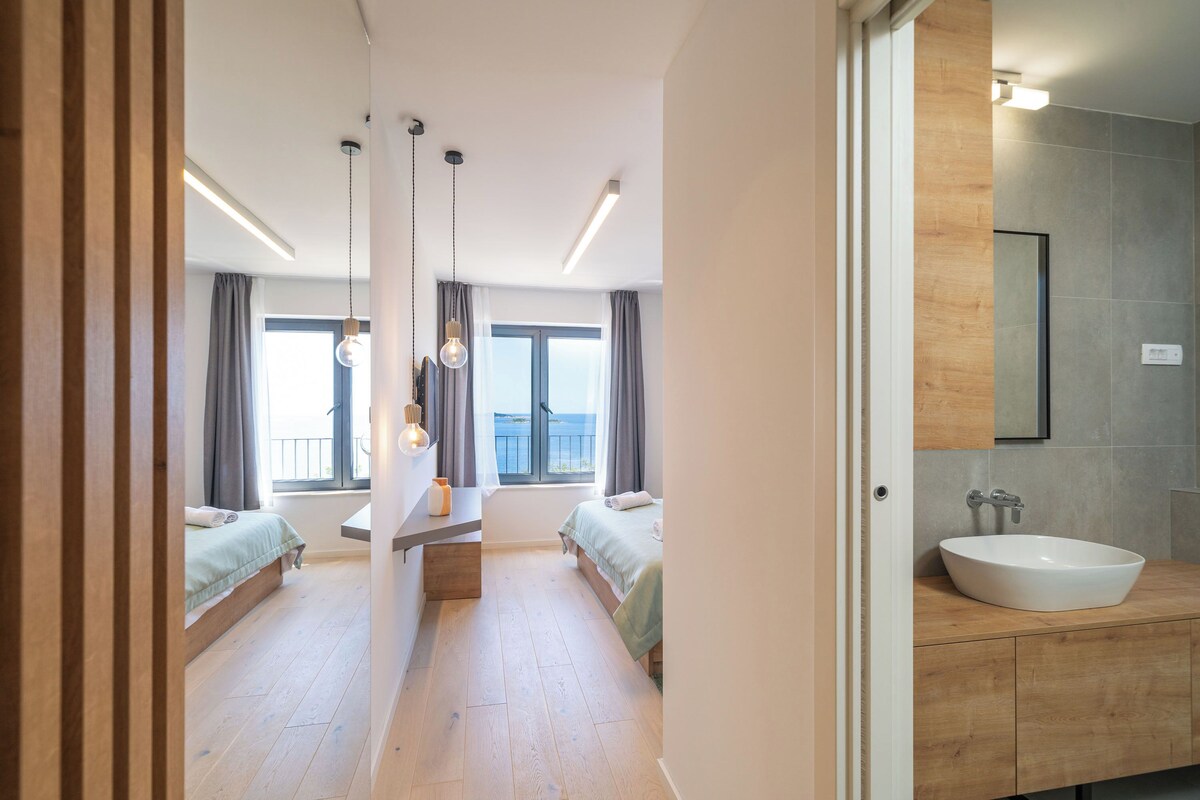 A modern hotel room with a large bed, a bedside table, a bathroom with a sink, and a view of the sea through the window.