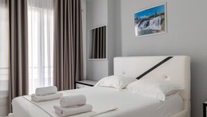 Una cama bien tendida con sábanas y toallas blancas, una mesita de noche, un espejo de pared y un cuadro con una cascada.