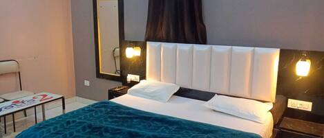 A hotel room with a bed, bedside tables, a small table with a chair, a mirror, and a window with curtains.
