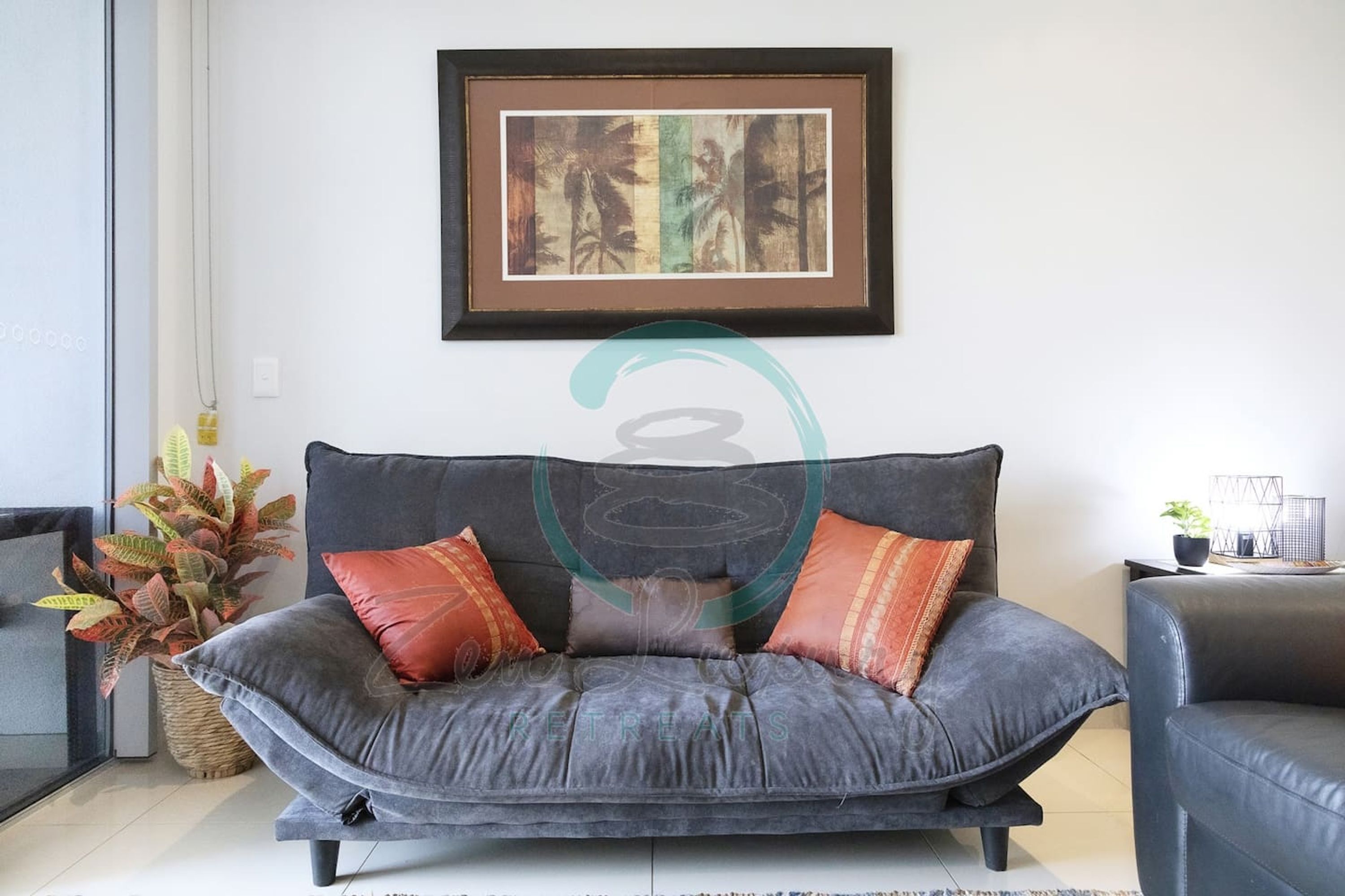 A grey sofa with orange cushions, a framed artwork above it, and a potted plant to the left.