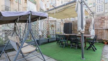Une terrasse avec un fauteuil suspendu, une table et des chaises. On y trouve également un téléviseur posé sur un meuble et une plante en pot.
