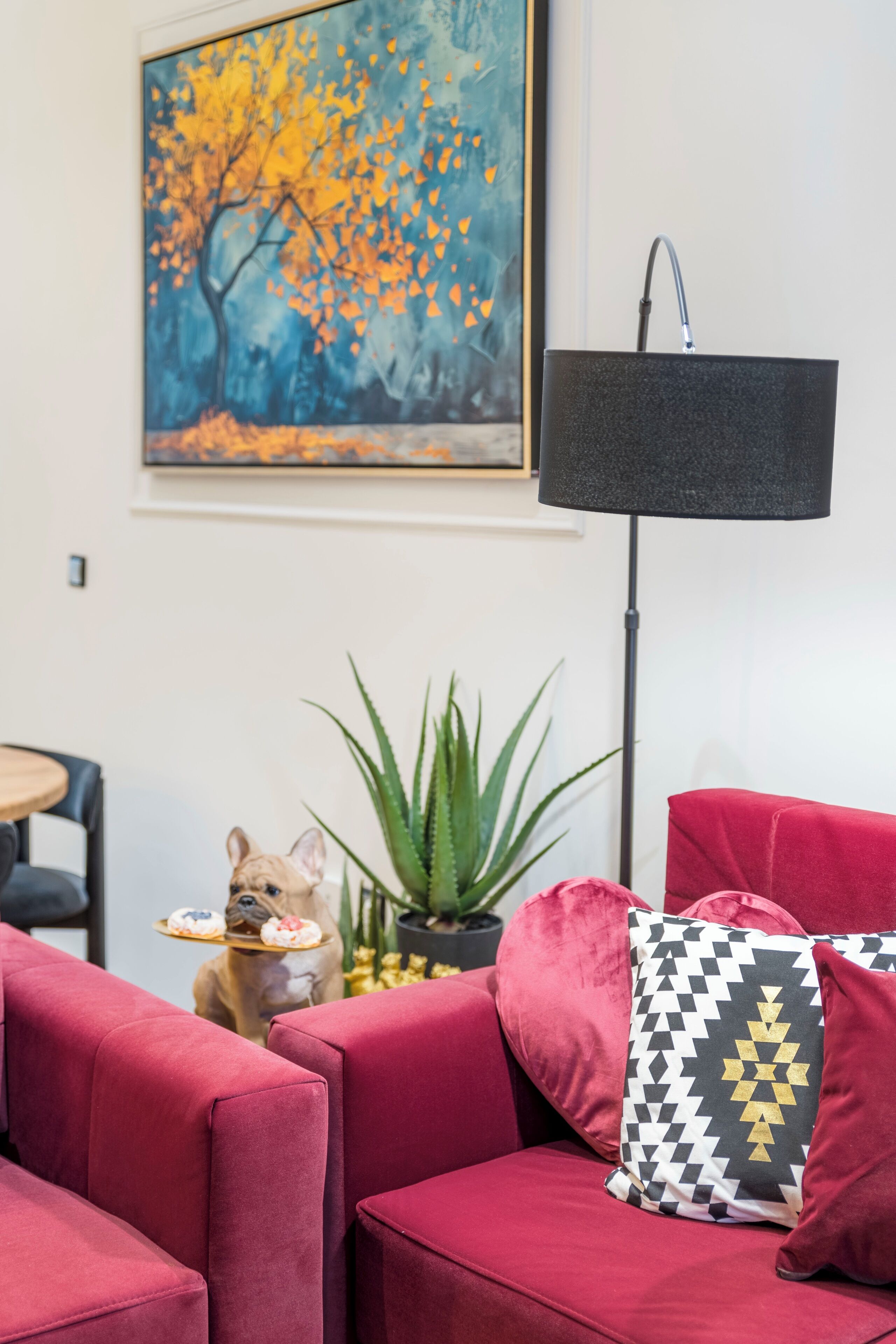Un canapé rouge avec un coussin à motifs noir et blanc, un lampadaire, un tableau représentant un arbre et un chien tenant un plateau.