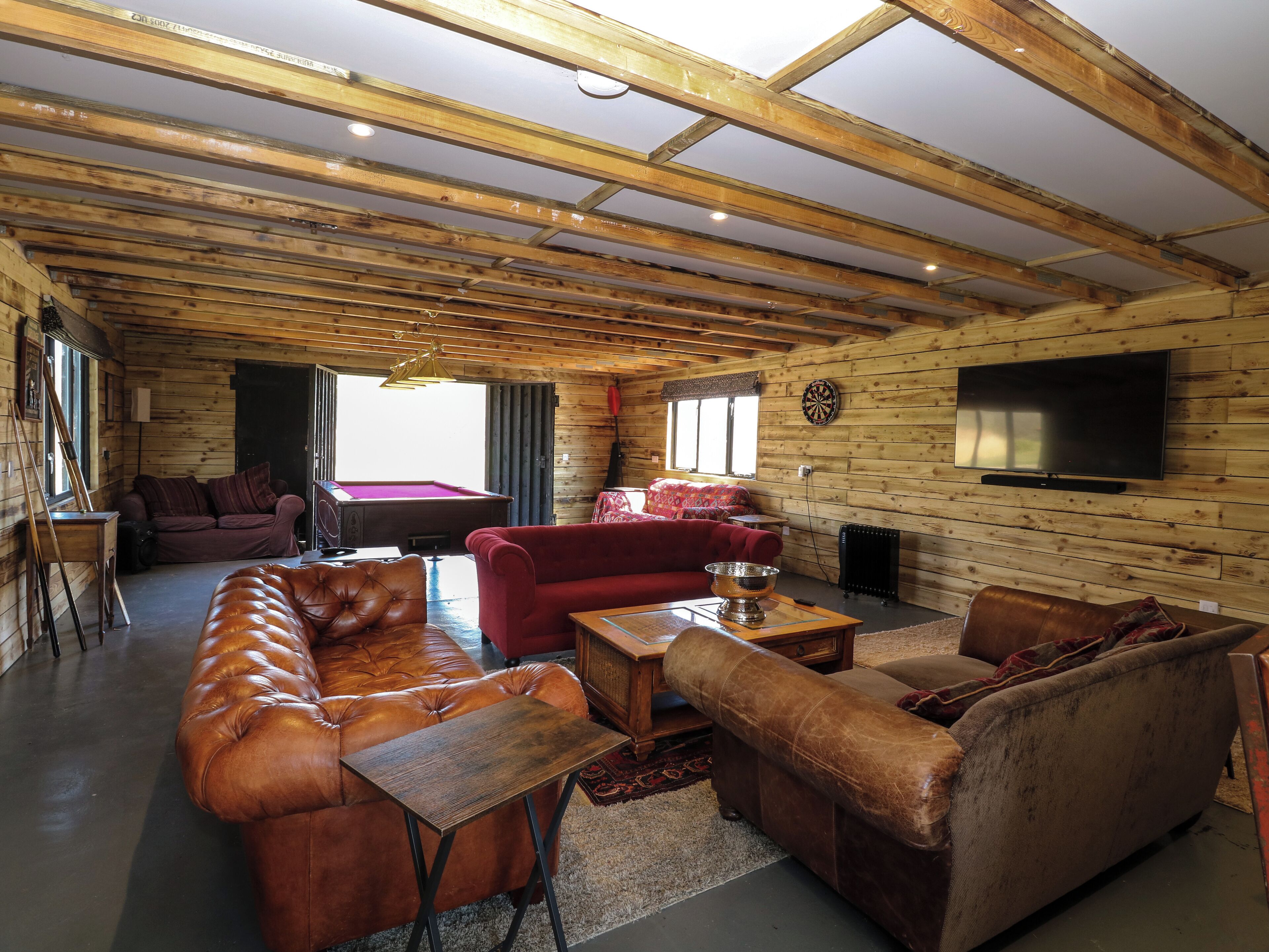 A log cabin interior with a wooden ceiling, a billiards table, leather sofas, and a flat-screen TV mounted on the wall.
