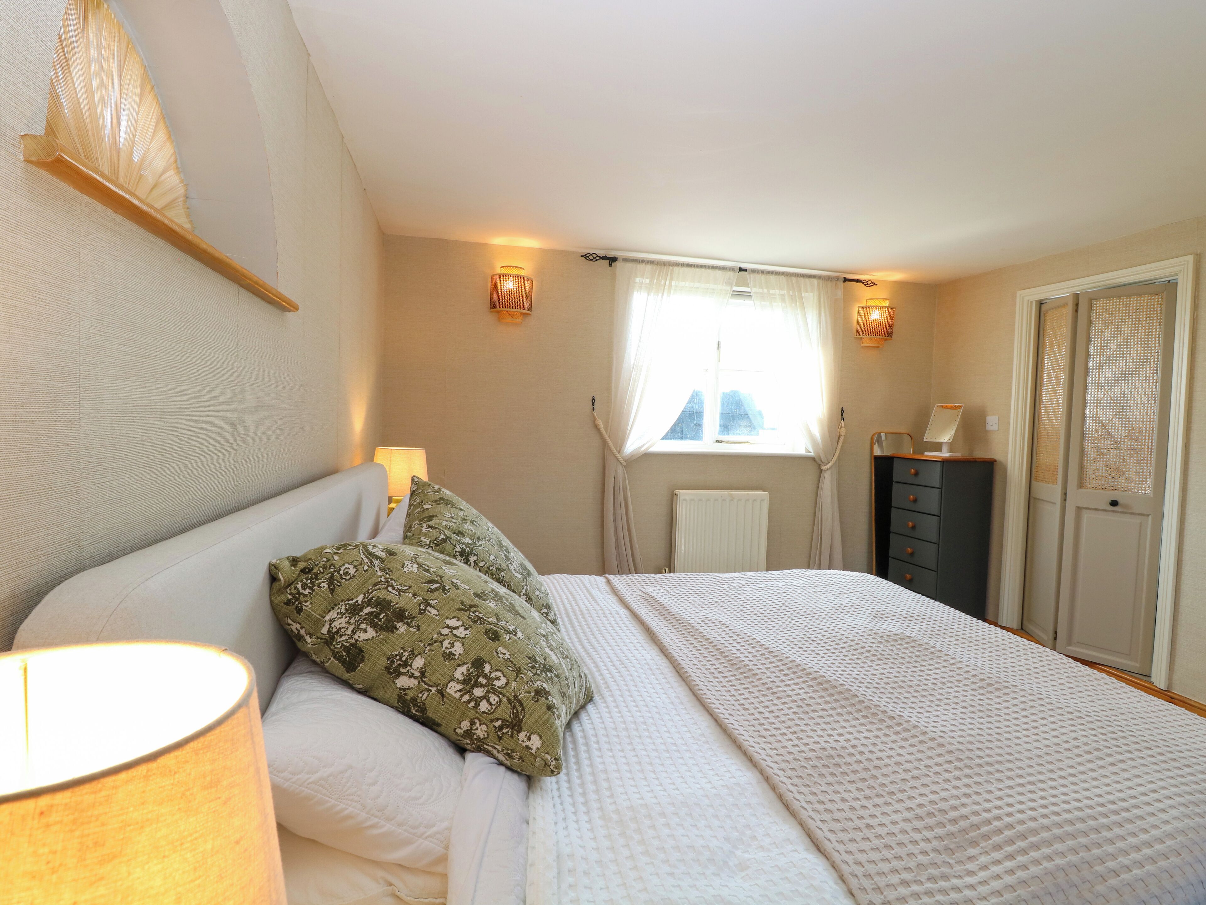 A bedroom with a bed, a nightstand, a lamp, a window with curtains, a radiator, and a wardrobe.