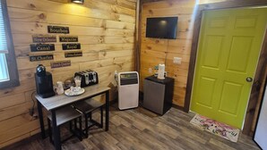 A cozy room with wooden walls and a green door, featuring a table with a coffee maker, a small fridge, and a TV mounted on the wall.