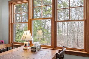 A dining area with a table, chairs, a lamp, and a tissue box. Large windows with a view of trees are visible.