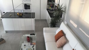 Eine moderne Küche mit weißen Schränken, einer schwarzen Arbeitsplatte und einem eingebauten Ofen. Im Wohnbereich steht ein Glastisch mit diversen Gegenständen darauf sowie ein weißes Sofa mit orangefarbenen Kissen.