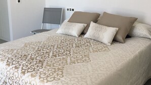 Ein ordentlich bezogenes Bett mit einem gemusterten Bettbezug, zwei Kissen und ein Stuhl in einem schlichten Zimmer mit weißen Wänden und Fliesenboden.
