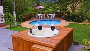 A hot tub with steps, surrounded by a wooden deck, with a pool and outdoor furniture in the background.