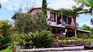 A house with a balcony, surrounded by lush greenery and a wooden bridge.
