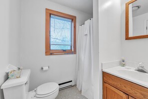 Et badeværelse med toilet, et vindue med persienner, bruseforhæng, vask og et spejl.
