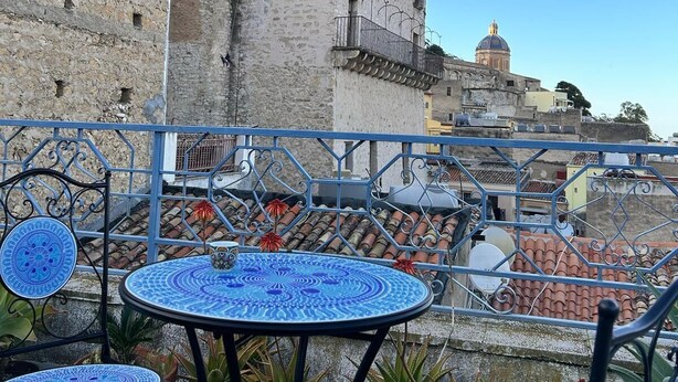 A terrace with blue-tiled tables and chairs, potted plants, and a view of a historic stone building with a tower. - Casa Di Sylwia (Termini Imerese)