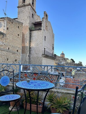 Une terrasse dotée de tables et de chaises aux carreaux bleus, de plantes en pot et offrant une vue sur un bâtiment historique en pierre, agrémenté d’une tour.