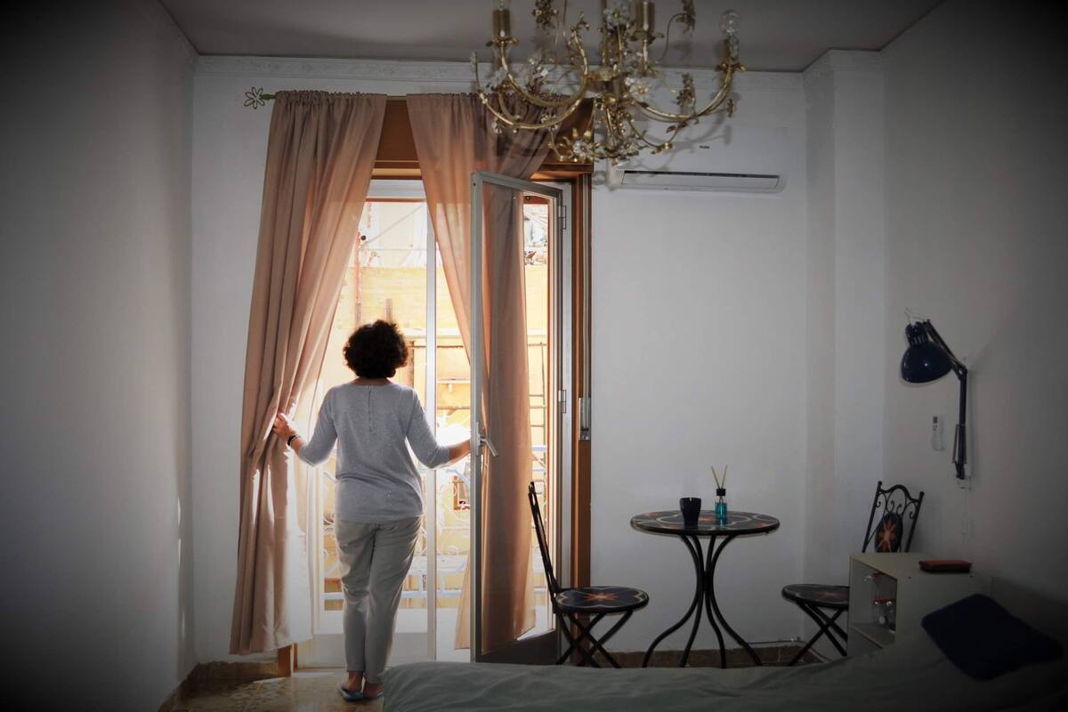 A room with a bed, a small table with a chair, a chandelier, and a window with curtains.