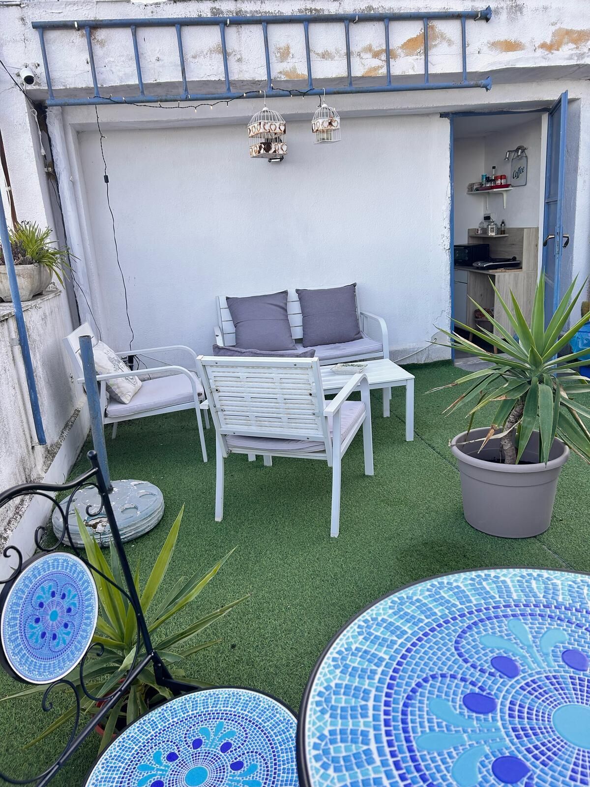 A small outdoor patio with a white sofa, a dining table with blue mosaic tiles, and a potted plant.