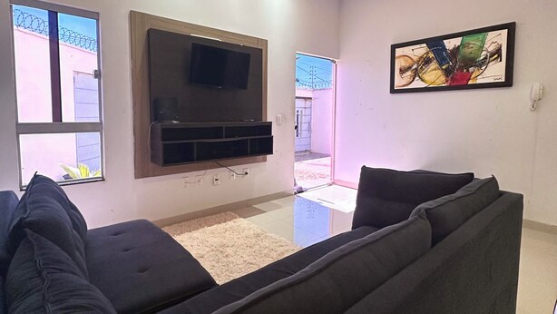 A modern living room with a large flat-screen TV mounted on the wall, a dark-colored sofa, and a small rug on the floor. - Casa Davisis 2  Jalapao e aeroporto (Palmas)