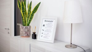 Ein gerahmtes Willkommensschild, ein Topfpflanze und eine Lampe auf einer weißen Kommode.