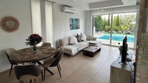 A modern living room with a dining area, a sofa, a TV, and a pool visible through the glass doors. - Aqua Luxe Ayia Napa Protaras Villa With Sea View (Paralimni)