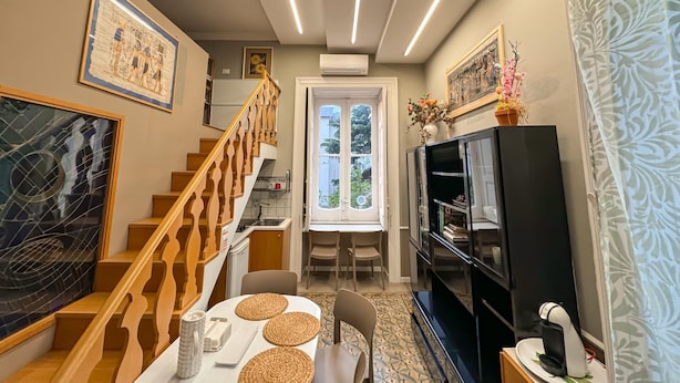A well-lit living room with a staircase, a dining area with a table and chairs, a black entertainment unit, and a large window with a view of greenery. - YourHome - Chiara's Apartment Sorrento 3 (Piano di Sorrento)