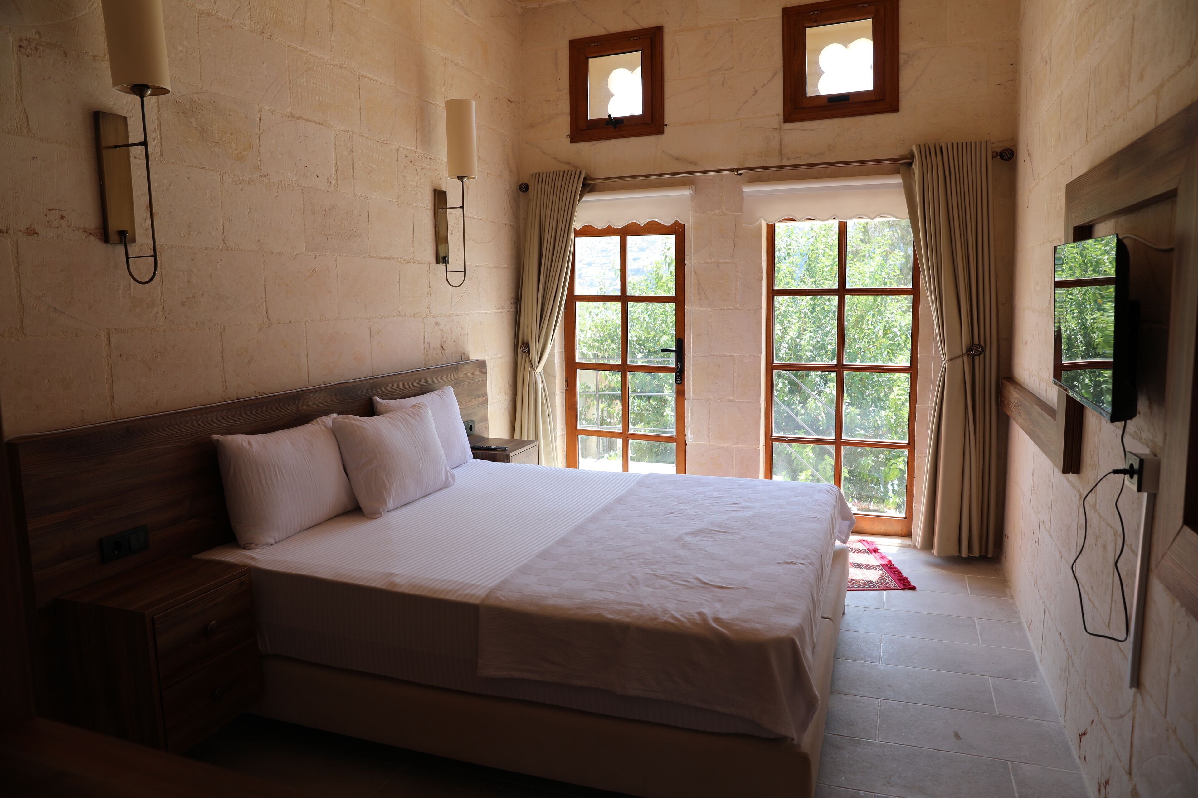 Une chambre à coucher avec un grand lit, une tête de lit en bois, deux tables de chevet, un tél éviseur fixé au mur et une fenêtre avec des rideaux.