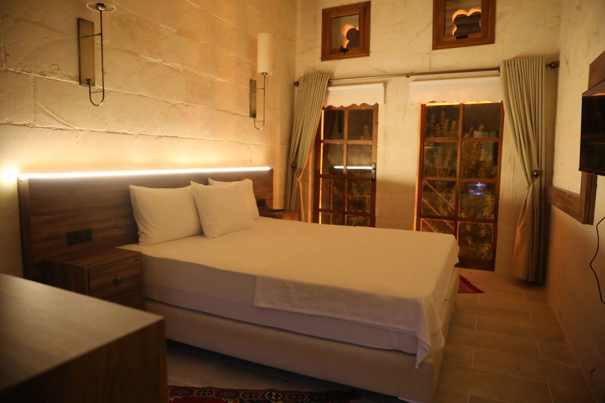 A bedroom with a large bed, wooden headboard, bedside tables, a nightstand, a wall-mounted lamp, and a TV.