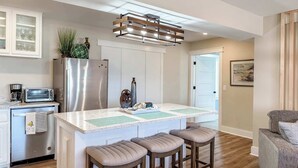 A modern kitchen with a central island, bar stools, a refrigerator, a microwave, and a hanging light fixture. - 7 Seas Ocean Breeze Sleeps Kitchens St George Island Pet Friendly Family (Eastpoint)