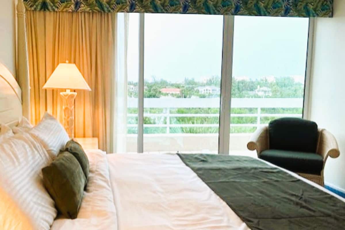 A hotel room with a bed, a nightstand with a lamp, a chair, and a view of greenery through large windows.