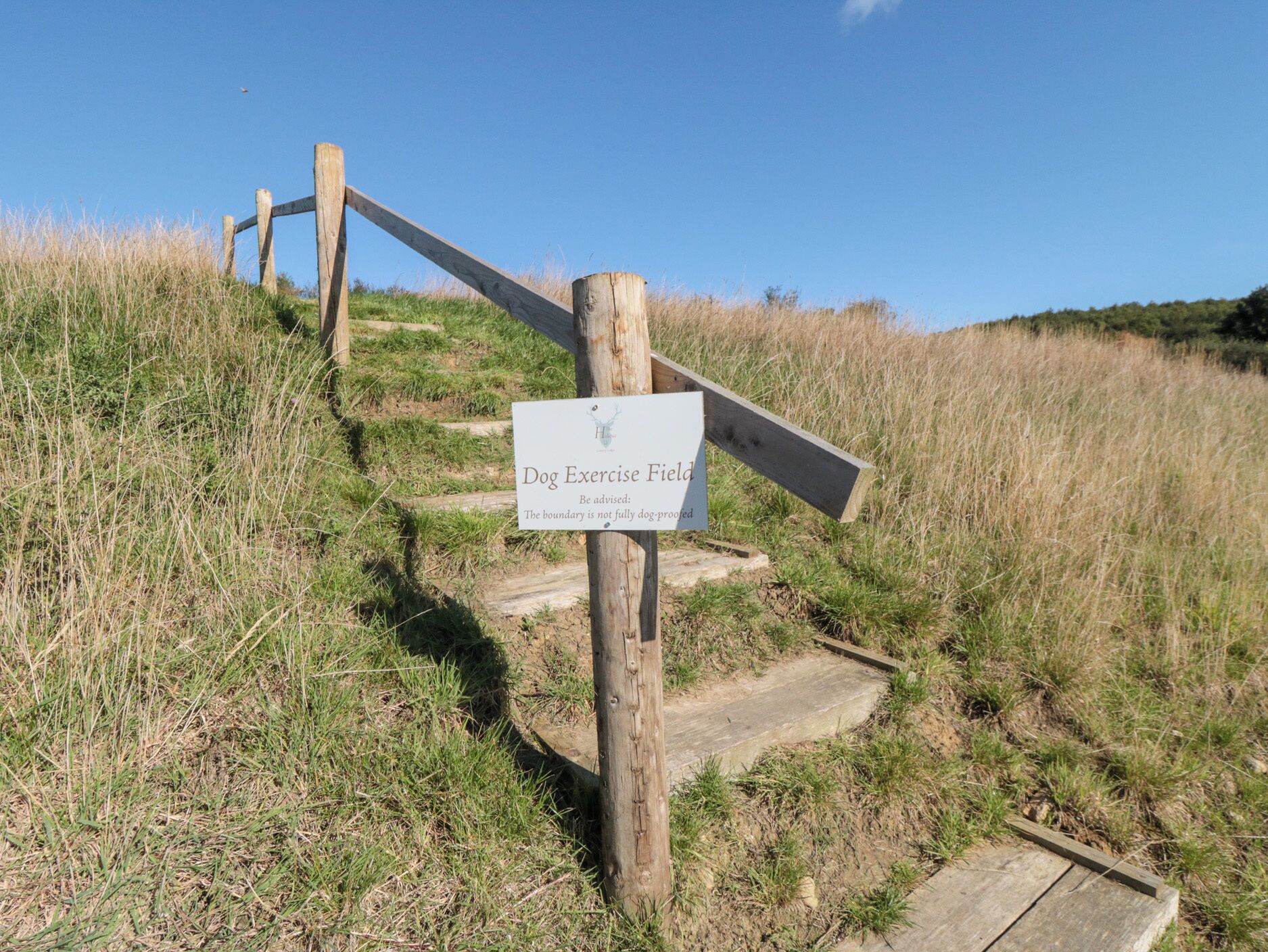 Un cartel de madera indica un campo para pasear perros, advirtiendo que el vallado no es completamente seguro.