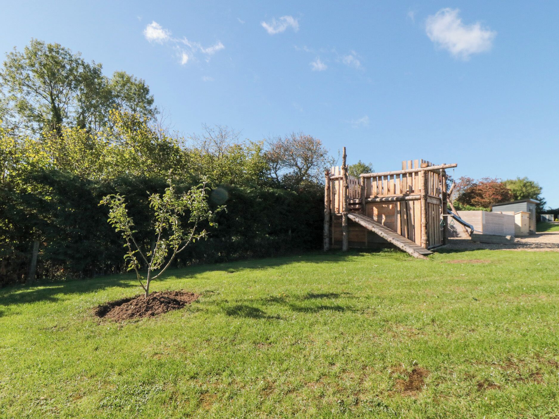 A wooden play structure with a slide and climbing area, surrounded by a grassy field and trees.